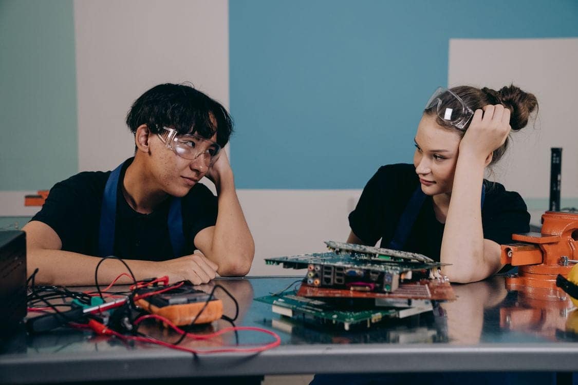 Two students in safety glasses leaning over a workbench covered with circuit boards, wiring and a multimeter